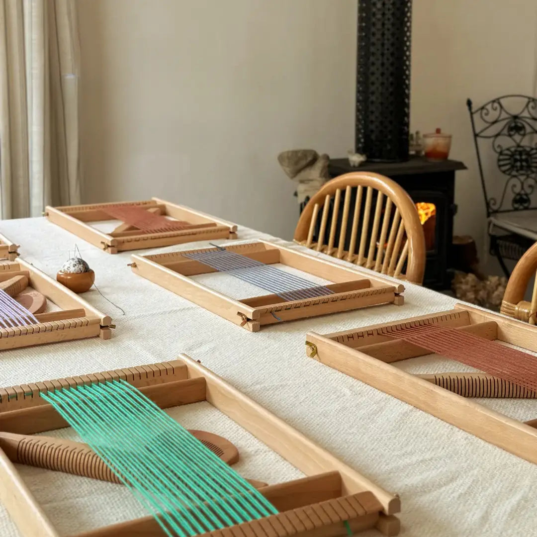 Several portable wooden frame weaving looms arranged neatly on a light-textured tablecloth. Each loom is warped with different colored threads, including vibrant green, slate blue, and terracotta. In the warm background, a cozy fireplace glows softly near wooden chairs, creating an inviting and professional atmosphere for a textile art workshop or creative session.