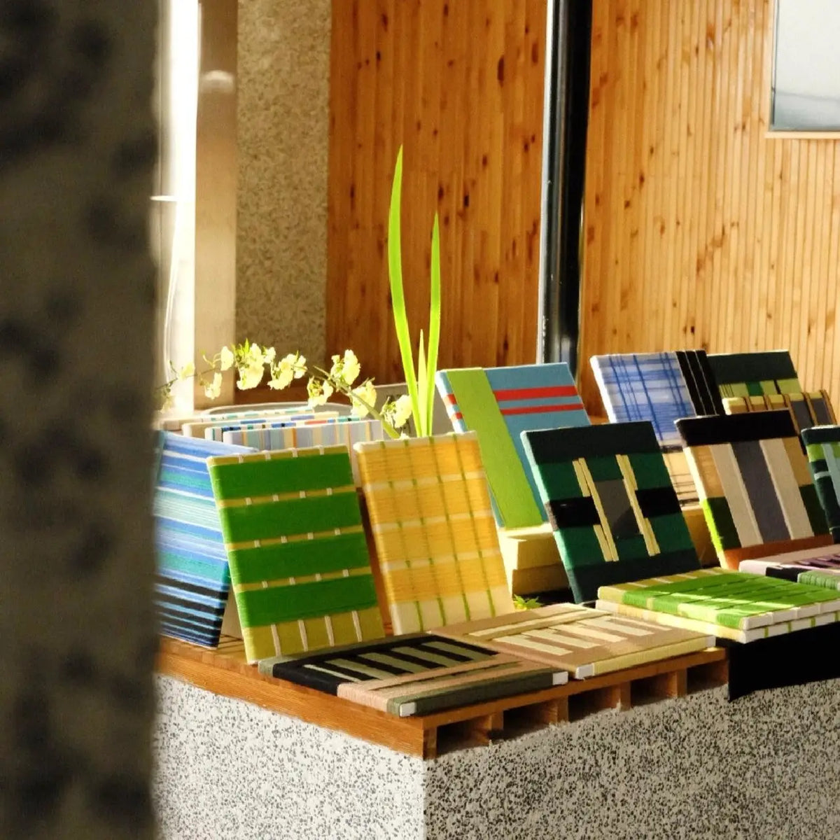 A beautiful gallery display of our premium handcrafted textile art swatches inside the Aassort studio. A variety of colorful, intricately woven fabric samples in vibrant green, bright yellow, and deep blues are lined up elegantly on a wooden display stand, set against a warm wooden wall backdrop.
