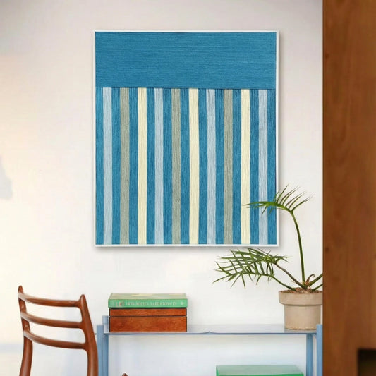 Blue Night blue and gray striped fiber wall art displayed above a console with books, a potted plant and a wooden chair