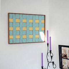 Horizontal textured wool wall art in a wooden frame placed on a white wall above a decorative candelabra with purple candles.