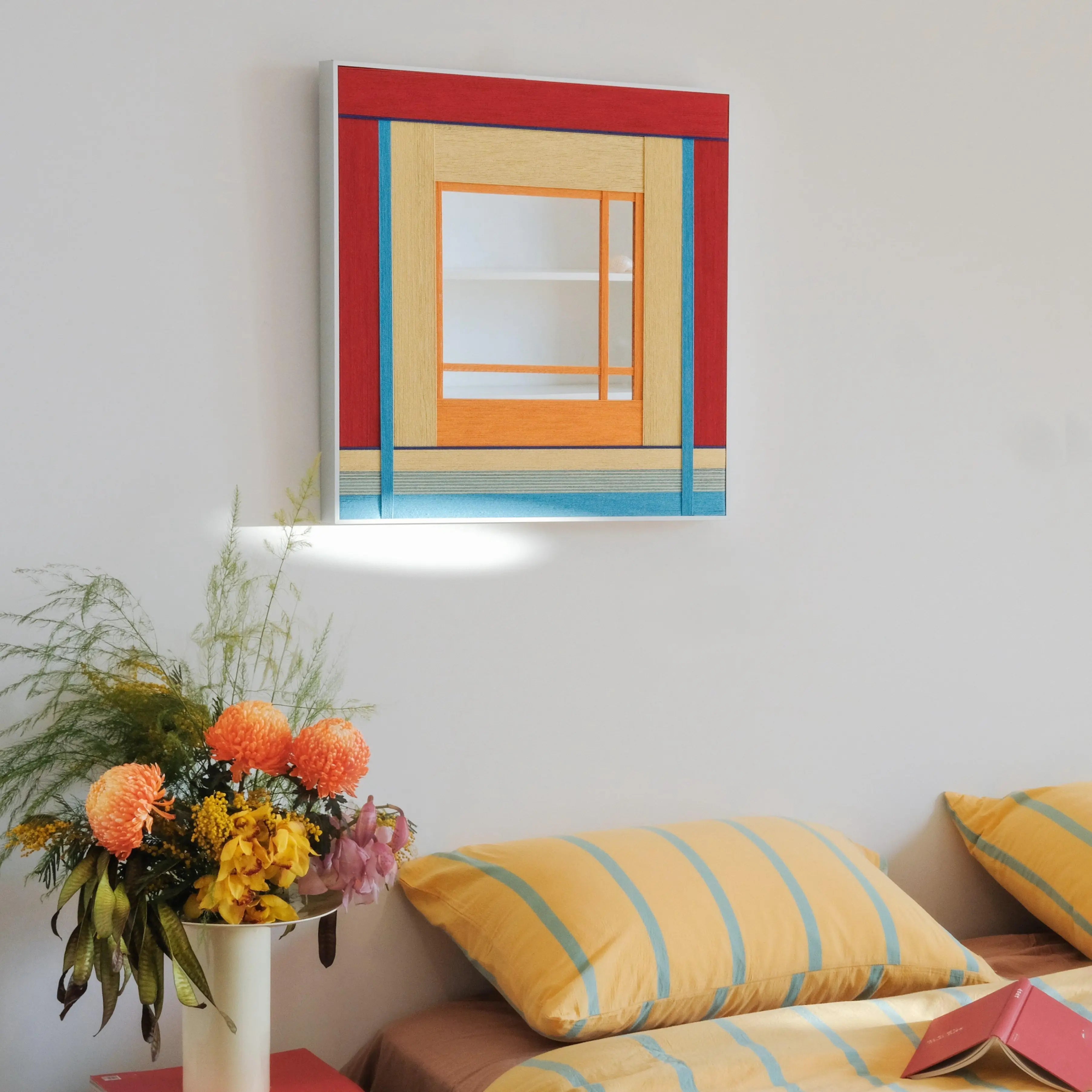 The geometric fiber art mirror styled in a bedroom setting above a bed with striped linens, creating a cohesive and cheerful aesthetic.