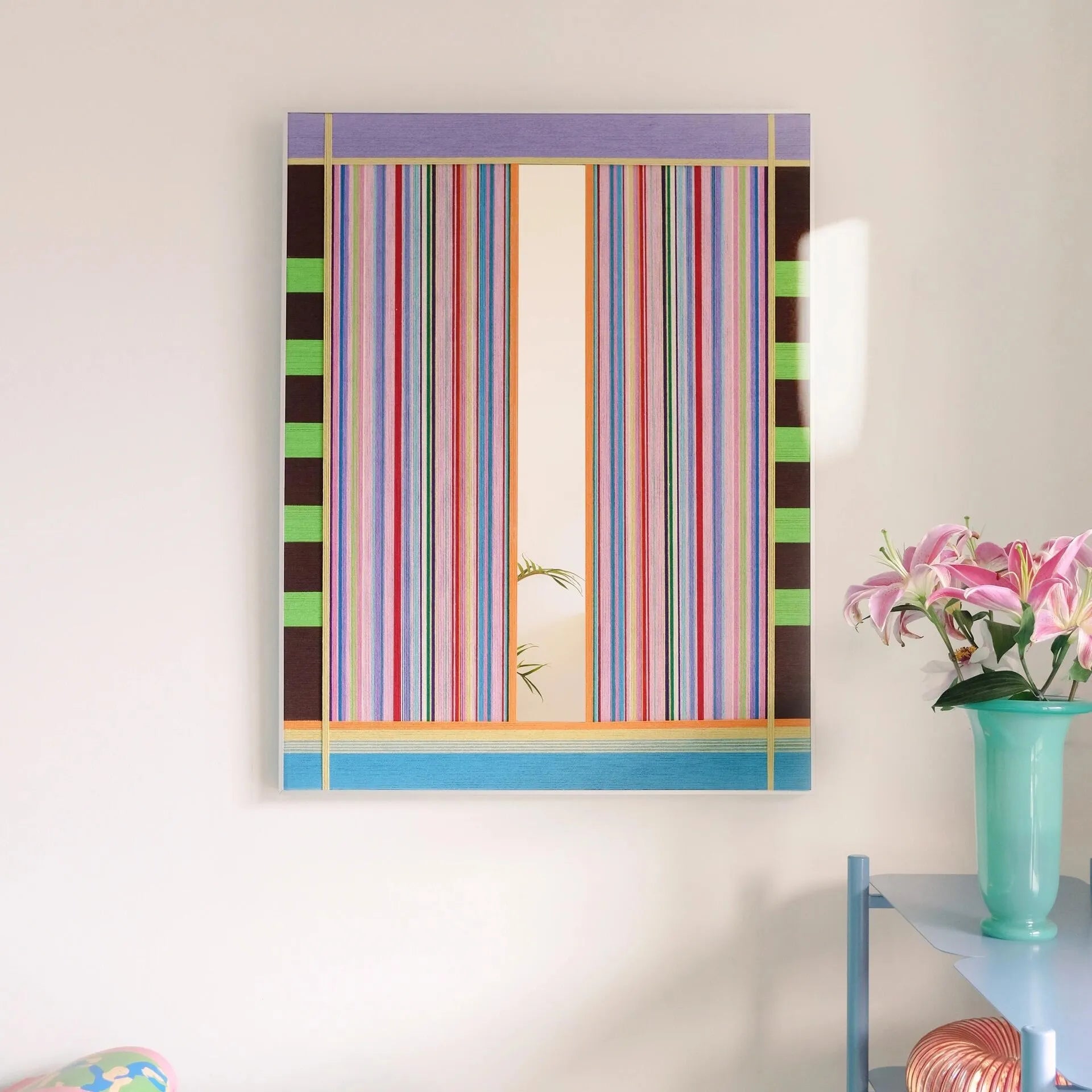Blooming Flowers hand-woven wool yarn wall art featuring alternating pastel pink, coral, mint, and sky-blue vertical stripes in a natural wood frame, displayed above a console table with vase and books.
