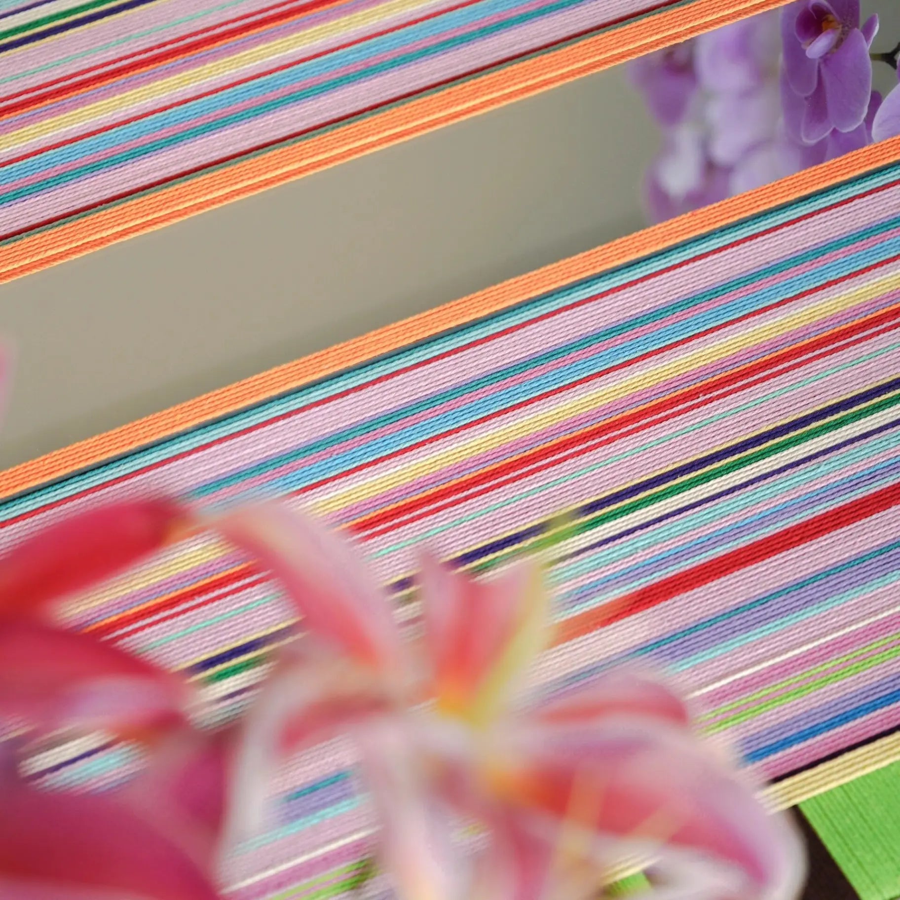 Hand-held view of Blooming Flowers yarn wall art in front of greenery, emphasizing its compact square form, rich looped texture, and multicolor stripe interplay.
