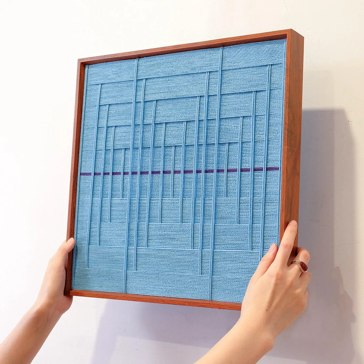 Hand-held view of Blue Bridge Over the Pond yarn wall art showing compact square form and precise grid-stripe pattern of soothing blue tones

