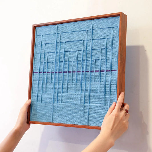 Hand-held view of Blue Bridge Over the Pond yarn wall art showing compact square form and precise grid-stripe pattern of soothing blue tones
