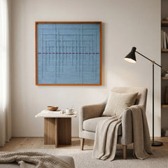 Framed light blue textile art displayed in a cozy reading corner above a light beige armchair and a modern black floor lamp.