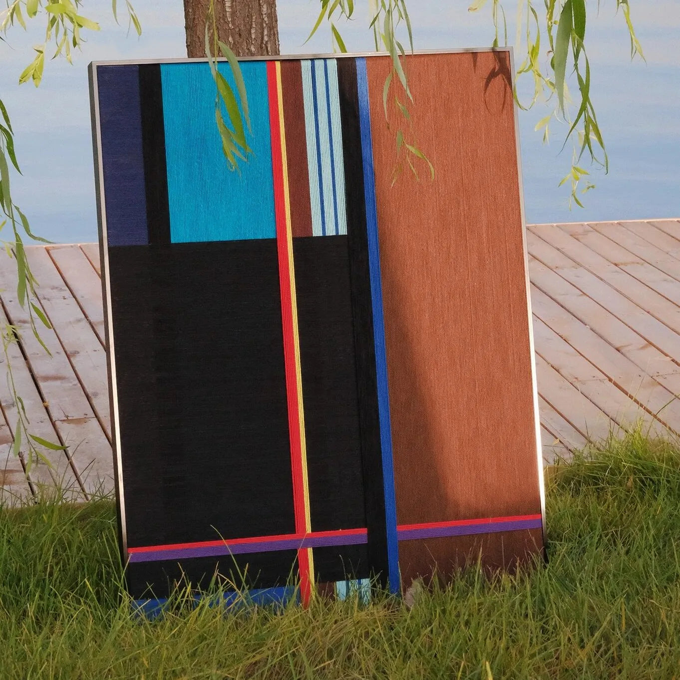 Blue Tide Night Run yarn wall art placed on a grassy riverbank, emphasizing its outdoor-friendly wool texture and vibrant stripe contrast.
