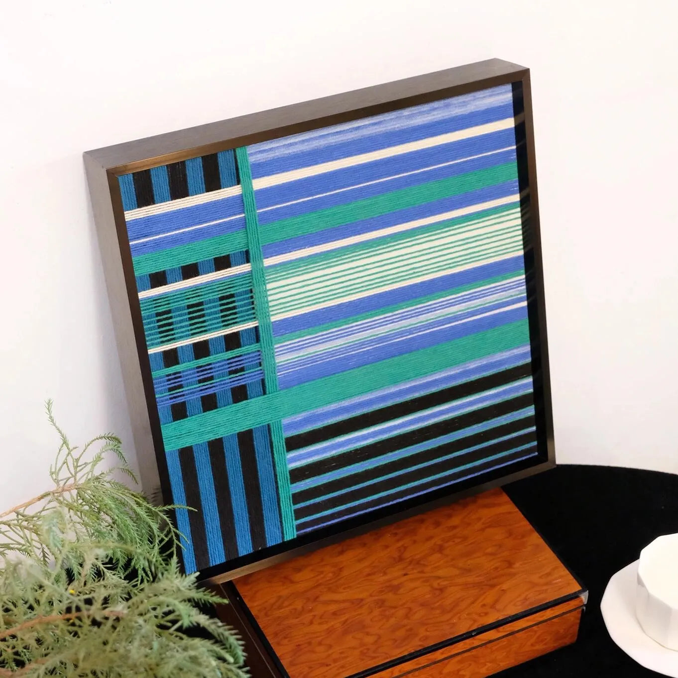Blues Harmony hand-woven wall art resting beside a laptop, showcasing its compact square format and harmonious blue-grey stripe pattern
