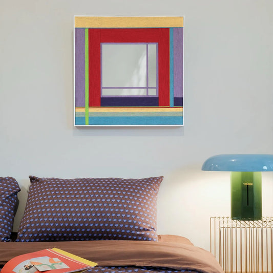 Bohemian Reverie hand‑woven yarn mirror wall art with pastel stripe borders and a deep red frame around a central reflective panel, displayed above a desk lamp and flowers.
