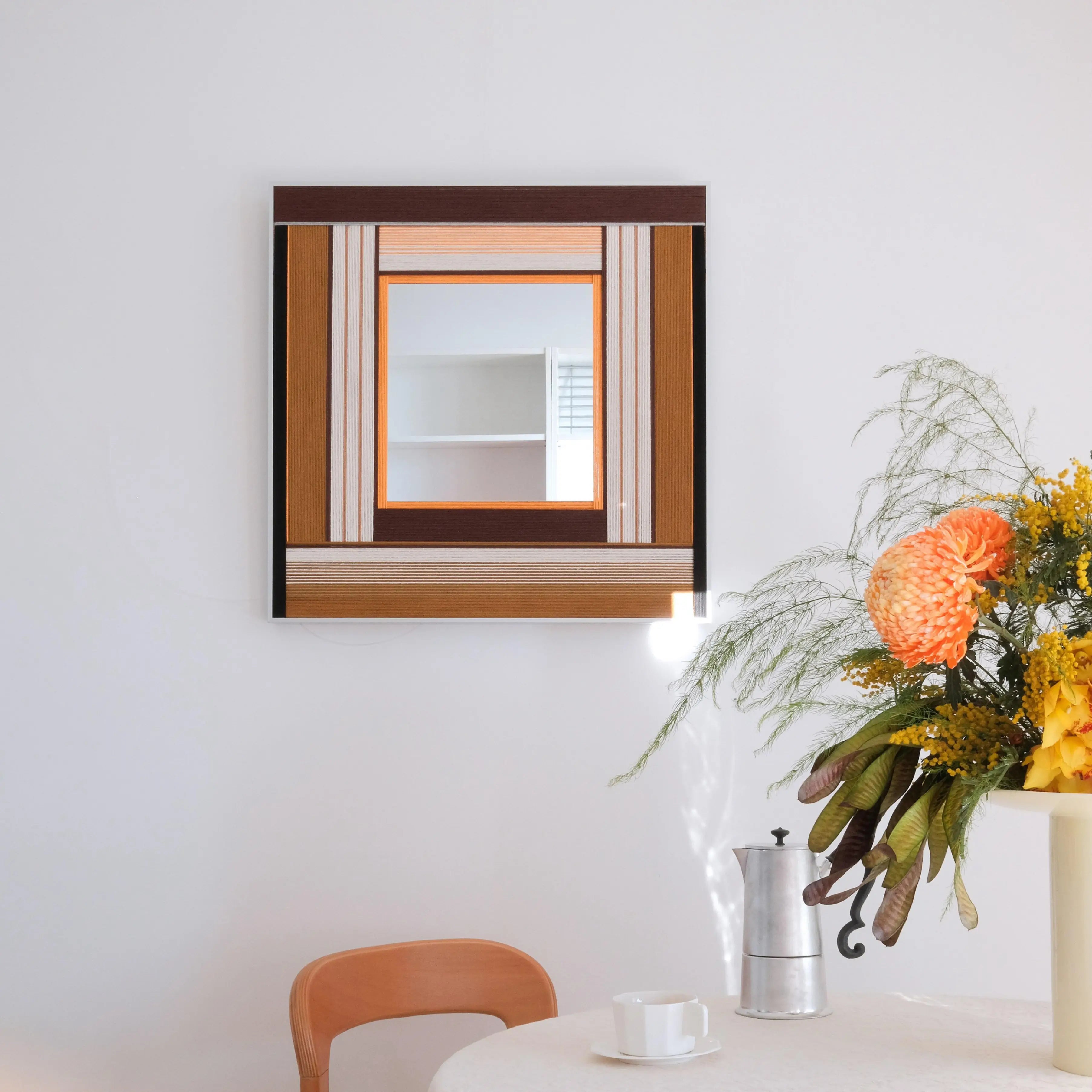 Framed hand-woven fiber art mirror titled Bread & Coffee featuring warm brown and beige stripes, styled with fresh flowers and a coffee pot.