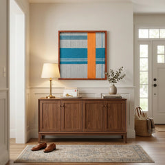 Framed geometric textile wall art displayed above a dark wood cabinet in a welcoming home entryway, softly illuminated by a table lamp.