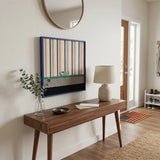 Framed striped and color-blocked textile art displayed above a dark wood console table in a minimalist entryway foyer.