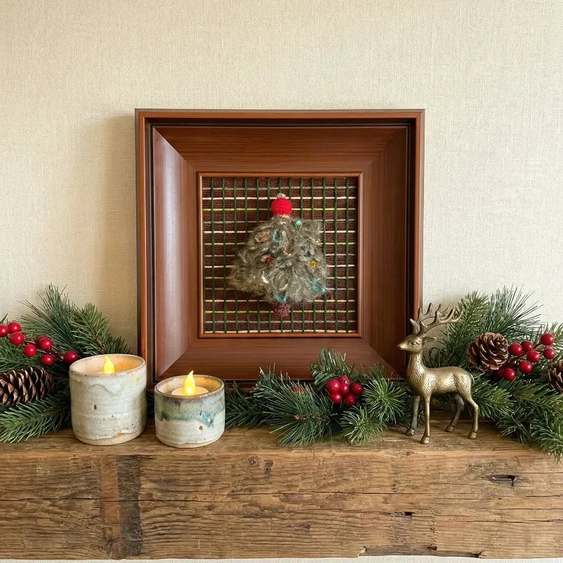 The textured fiber art piece resting on a rustic wooden surface, flanked by candles and pine branches for a cozy seasonal look.