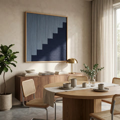 Framed fiber art tapestry with a geometric blue gradient design hanging near a wooden dining table and contemporary buffet cabinet.