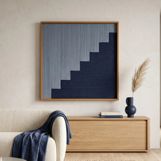 Square fiber wall art with a blue staircase motif placed above a minimalist wooden sideboard with dried botanical accents and a blue throw.
