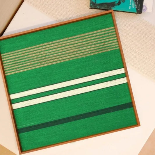 Endless Flow hand-woven wool yarn wall art with green stripe pattern displayed on a desk alongside yarn balls and a lamp, highlighting its square format and vibrant texture
