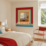 Fields of Blossoms red, blue, cream, and orange yarn-framed mirror displayed above a bed in a sunlit modern bedroom.