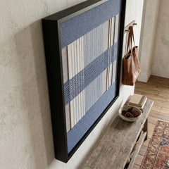 Angled side view of the framed Foggy Pier textile wall art hanging on a textured wall, showing the black frame's depth above a rustic wooden entryway bench.
