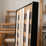 Angled close-up of the Forgotten Gateway hand-woven wool tapestry featuring cream and dark brown stripes set within a sleek black frame.