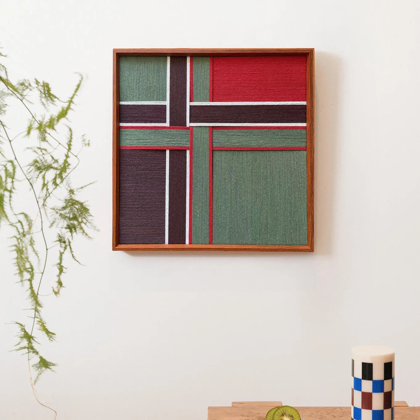Framed hand-woven wool art featuring geometric red and green blocks with white lines resembling a gift ribbon, displayed on a white wall.