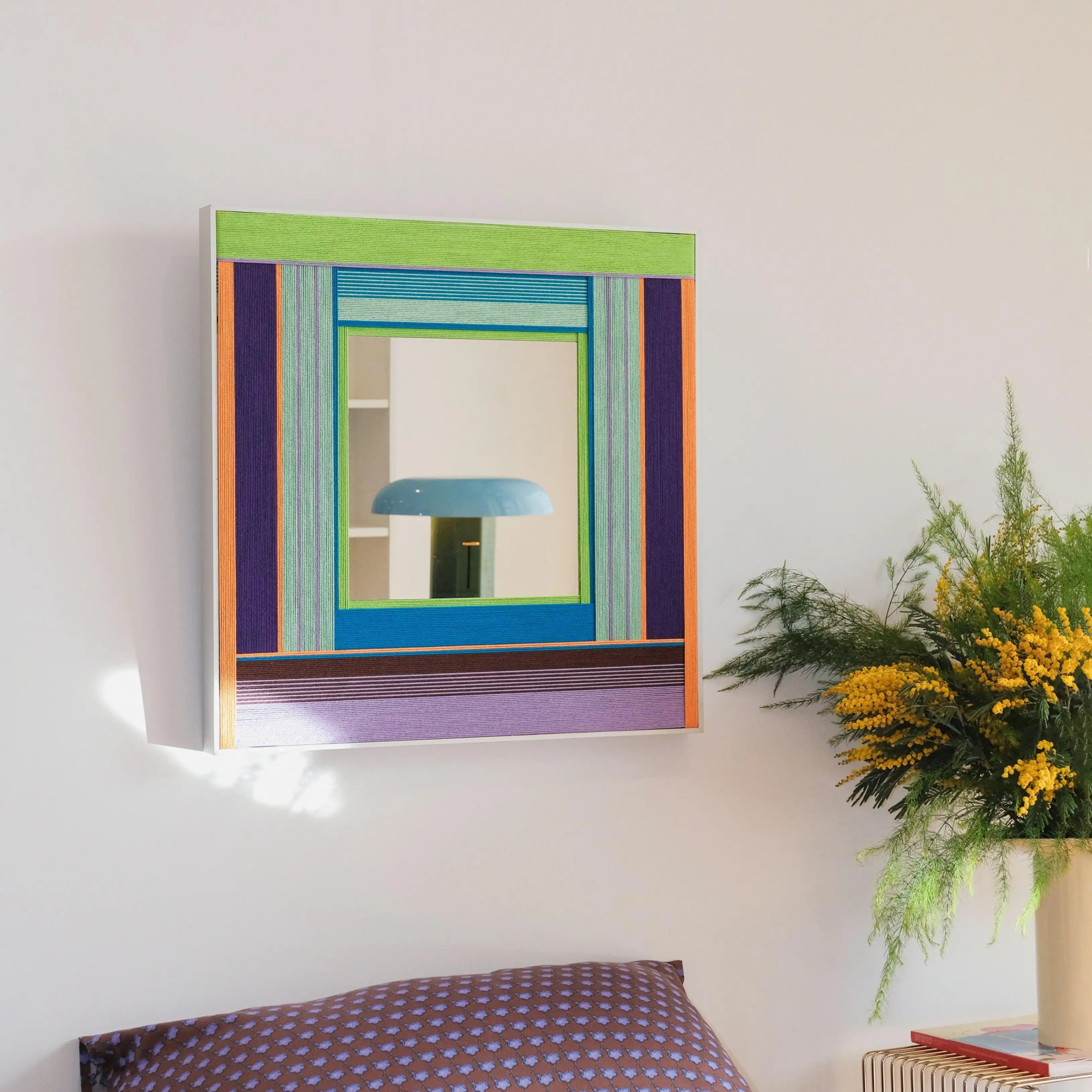 Hidden Forest mirror art mounted on a wall next to a potted plant, showcasing its deep green and purple stripe pattern around the reflective center.
