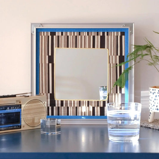 Inception hand-woven wool yarn wall art with integrated square mirror center and alternating oatmeal and sky-blue stripe panels in a natural wood frame, showcased on a tabletop
