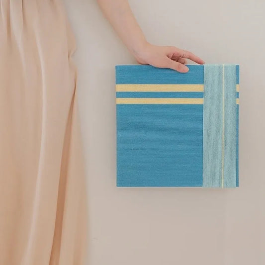 Infinite Serenity hand-woven wool yarn wall art featuring two narrow pale yellow horizontal stripes above a serene blue panel with a vertical mint stripe, all framed in natural wood
