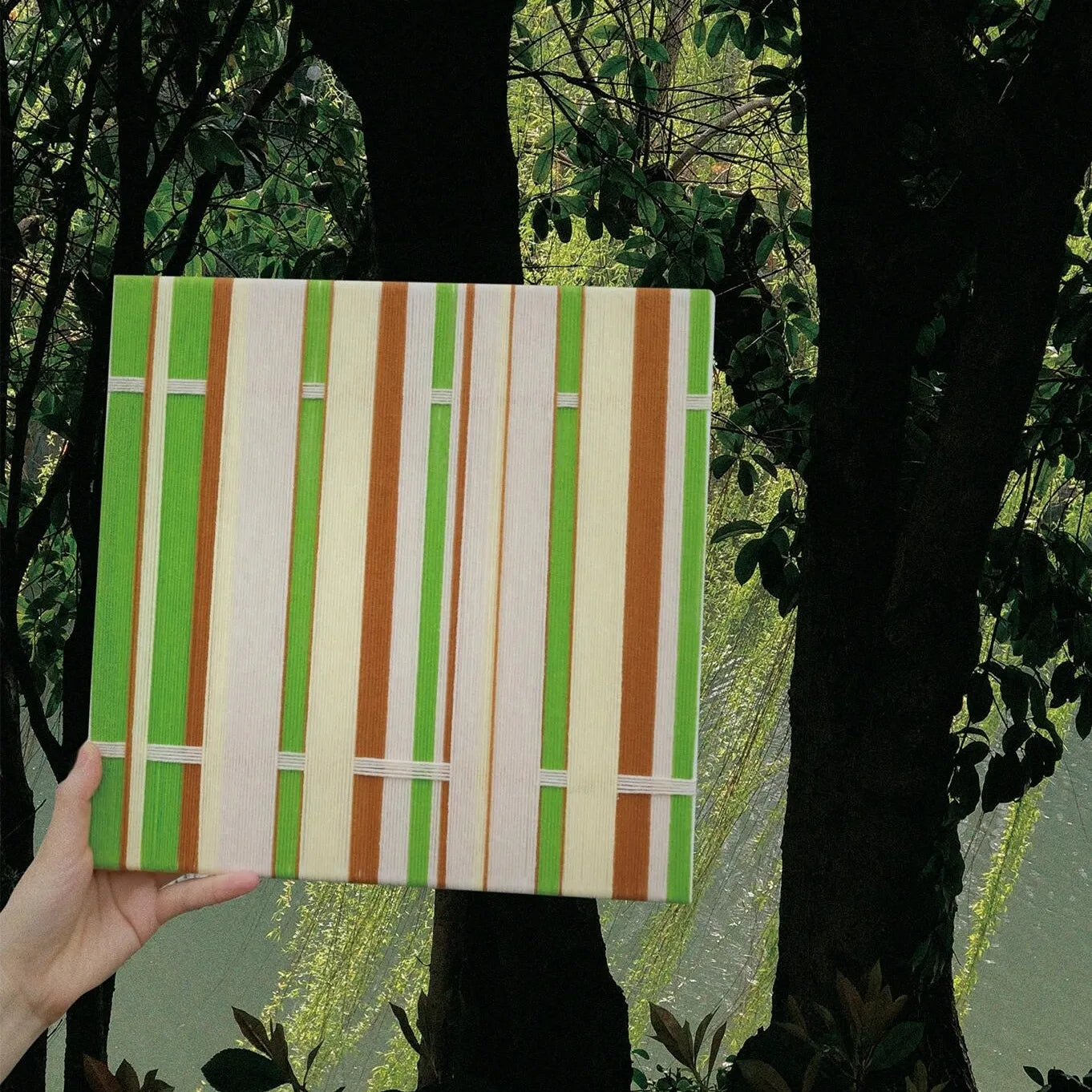 Hand-held view of March Tree yarn wall art in front of leafy trees, highlighting its sturdy wood frame and fresh green stripe palette.
