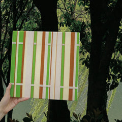 Hand-held view of March Tree yarn wall art in front of leafy trees, highlighting its sturdy wood frame and fresh green stripe palette.
