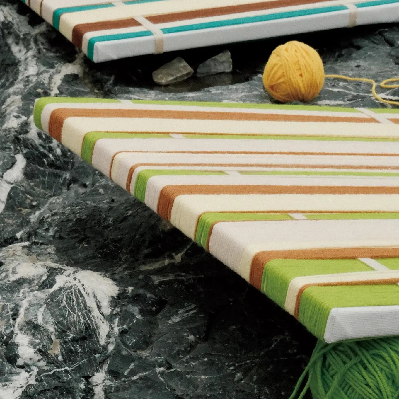 March Tree hand-woven yarn art resting on a stone bench outdoors, emphasizing its vibrant green and white stripes against a natural background.
