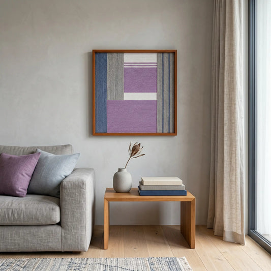Framed textured fiber wall art hanging in a bright contemporary living room above a wooden side table and grey sofa.