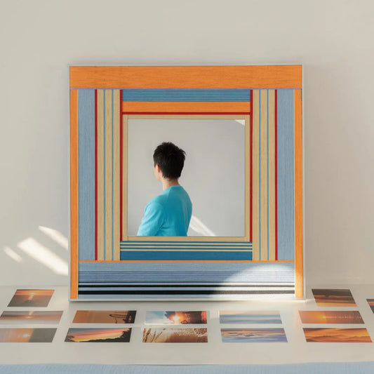 Square woven textile mirror reflecting a person in a blue shirt, styled resting on a table surrounded by scattered printed photographs in bright sunlight.