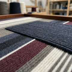 Extreme low-angle macro shot of the Midnight Fjord hand-woven textile art lying flat, highlighting the textured navy patch over burgundy, grey, and cream yarn stripes.