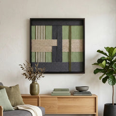 Large square fiber wall art in forest tones hanging above a light wood sideboard in a bright, minimalist living room.