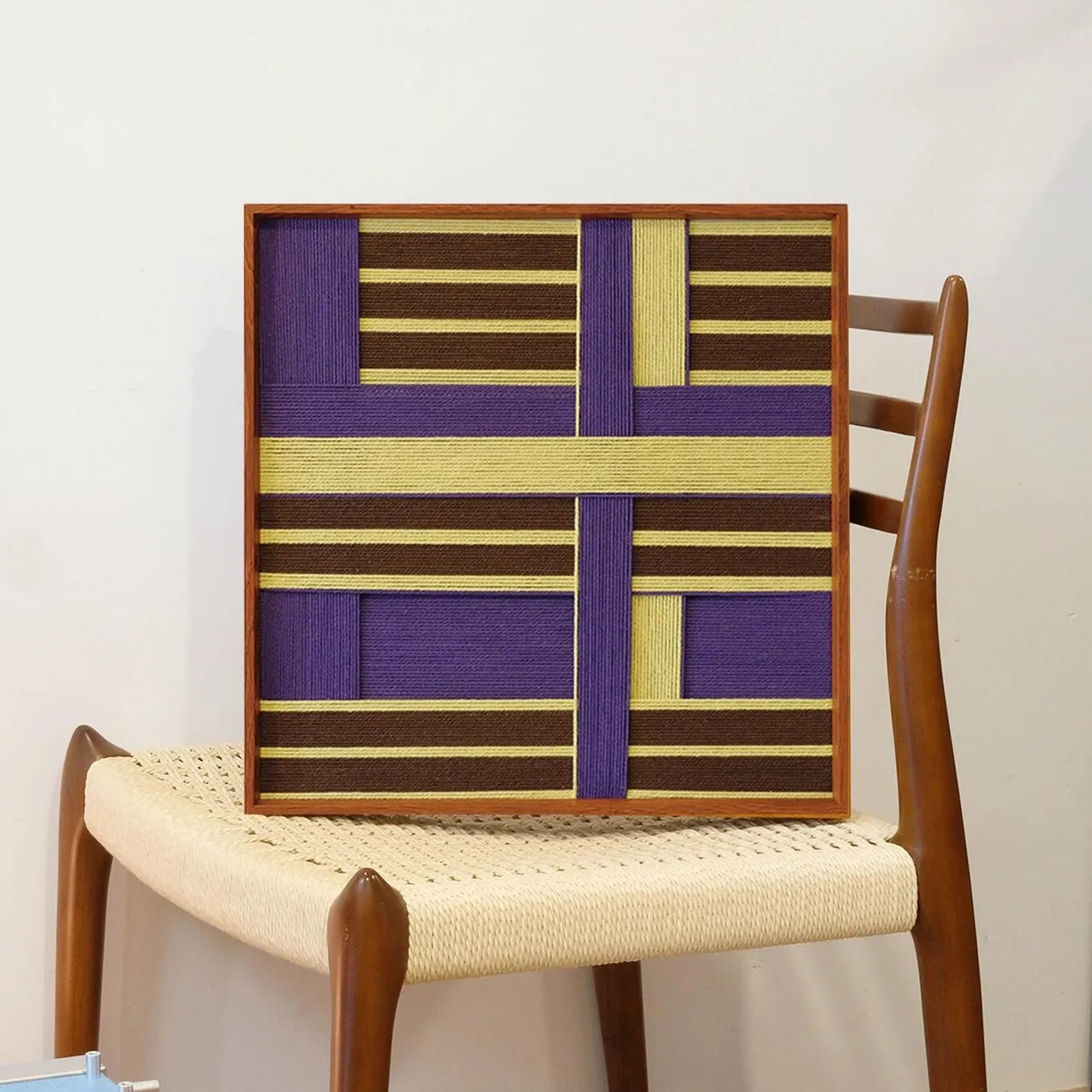 Mrs. Maisel yarn wall art leaning on a wooden chair, highlighting its precise grid pattern and sturdy frame.

