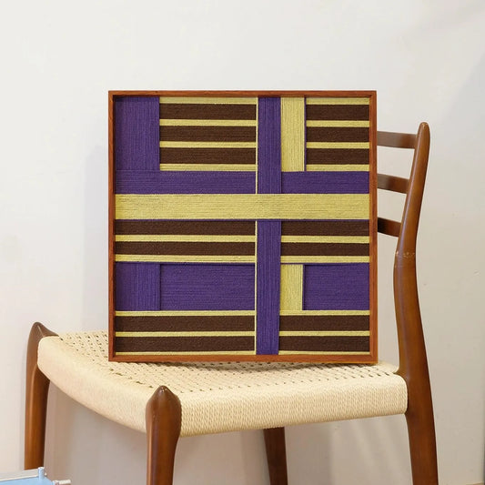 Mrs. Maisel yarn wall art leaning on a wooden chair, highlighting its precise grid pattern and sturdy frame.

