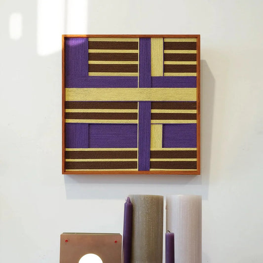 Mrs. Maisel hand-woven wool yarn wall art featuring alternating lavender and gold grid panels in a natural wood frame, displayed above a console table with decorative vases.
