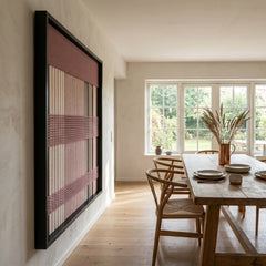 Striking square hand-woven wool tapestry positioned perfectly on a dining room wall, viewed from an angle overlooking a light wooden dining table and chairs next to large bright windows.