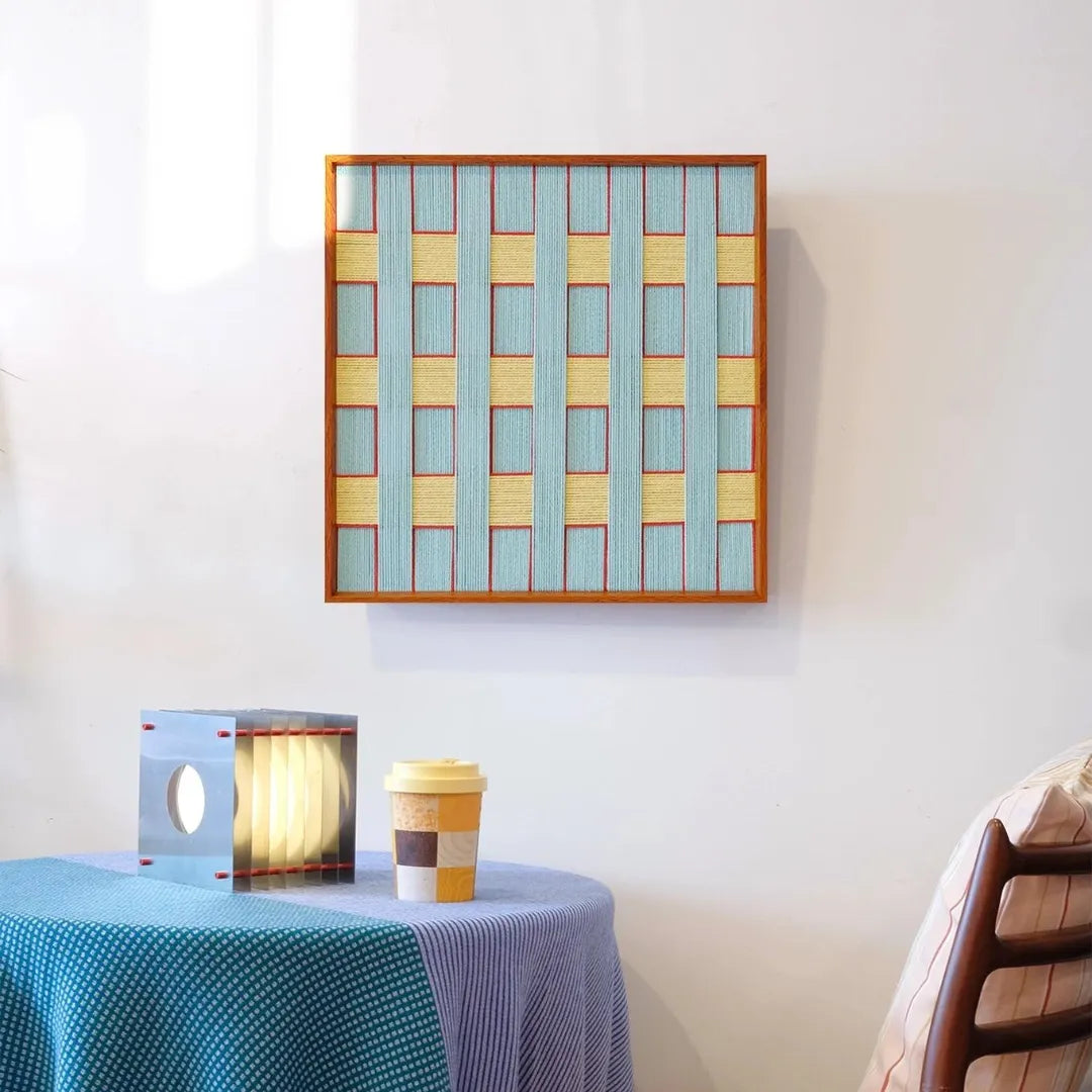Push Open the Afternoon Window wall art with alternating pastel blue and buttery yellow yarn stripes in a square grid pattern and natural wood frame
