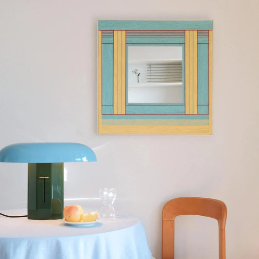 Pear Blossom Tree hand‑woven yarn mirror wall art featuring alternating spring‑green, mint, and natural wood stripe borders framing a central reflective panel, displayed above a desk with a teal lamp and potted flowers.
