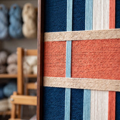 Close-up corner detail of the hand-woven wool tapestry highlighting textured vertical blue and cream yarns intersected by a salmon horizontal band in a wooden frame.