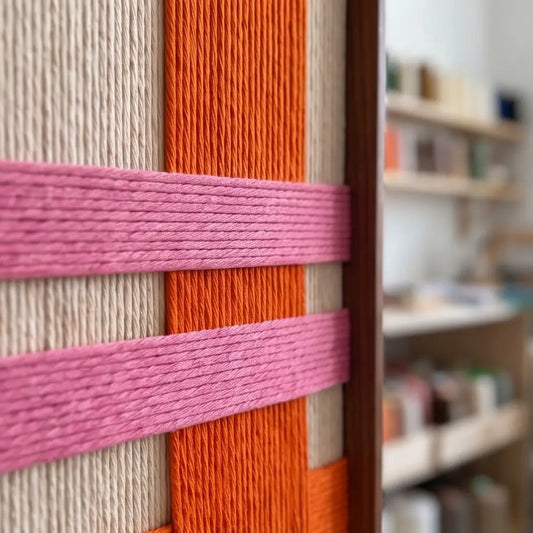 Close-up macro detail of the hand-woven wool tapestry showing textured vertical cream yarns intersected by bold orange and pink horizontal bands in a metal frame.