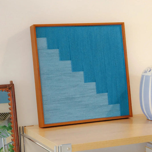 Radiant Azure hand-woven yarn wall art in square format with gradient blue shades, displayed on a wooden shelf for a serene coastal feel
