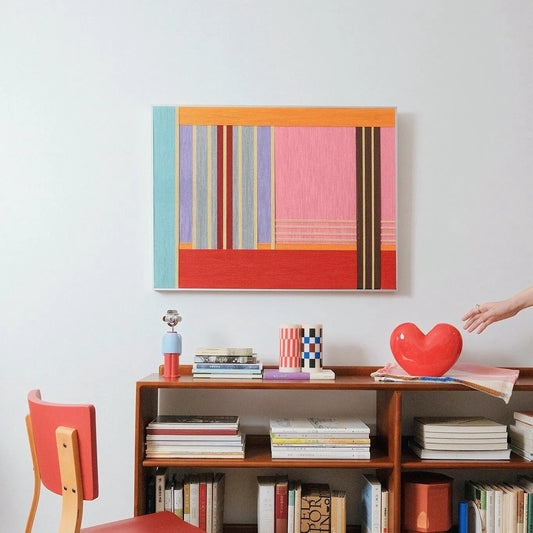 Rainbow Hearts wall art leaning on a tabletop beside a coffee cup, highlighting the bold red and pink stripe blocks and tight wool texture
