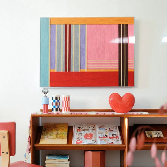 Rainbow Hearts hand-woven wool yarn wall art in a natural wood frame featuring vibrant red, pink, yellow, and blue stripes, displayed on a console shelf with a red heart ornament and mugs
