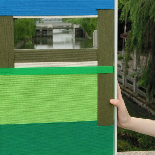Handheld view of Riverside RE006 textile mirror art showing its framed format, central mirror, and precise blue and green yarn bands.