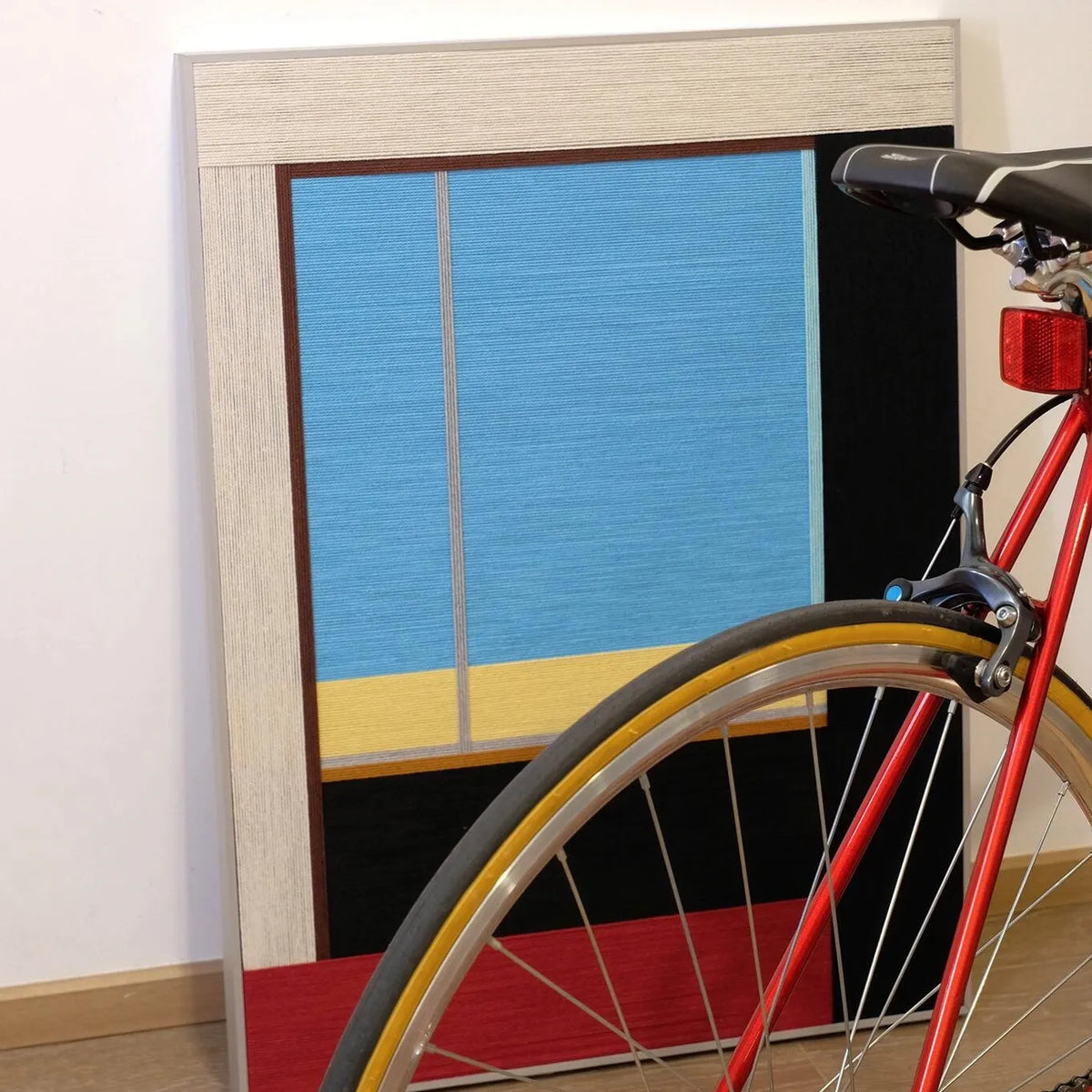 After the Beach Walk RE007 yarn wall art displayed beside a bicycle wheel, highlighting its vibrant blue, gold, and red striped composition.