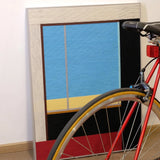 After the Beach Walk RE007 yarn wall art displayed beside a bicycle wheel, highlighting its vibrant blue, gold, and red striped composition.