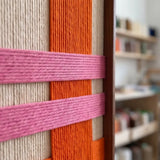 Macro detail of Pop Boulevard textured textile art showing cream vertical yarn crossed by bold orange and pink bands in a metal frame.