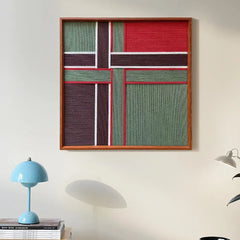 The Gift Ribbon textured wall hanging styled above a modern blue lamp, showcasing its vibrant red and green geometric pattern in a home setting.